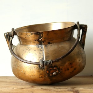 Op de afbeelding: Een vintage messing ketel met een afgeronde body en een brede opening. De ketel heeft een stevige handgreep en wordt ondersteund door een houten oppervlak. Het messing heeft een warme, verouderde patina, wat wijst op gebruik in de loop der tijd. De ketel wordt waarschijnlijk gebruikt om te koken of voor decoratieve doeleinden.