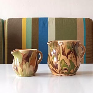 Puede incluir: Dos jarras de cerámica con un diseño marmoleado en tonos marrones, verdes y crema. La jarra más grande tiene un asa en el lado derecho, mientras que la jarra más pequeña tiene un asa en el lado izquierdo.