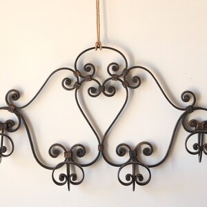 Op de afbeelding: Een zwart metalen wanddecoratief stuk met een gerold ontwerp en vijf haken. Het stuk hangt aan een bruin touwtje.