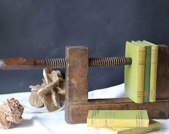 May include: A vintage wooden book press with a metal screw and handle. The press is holding a stack of books with green and yellow covers. The books have text on the spines.