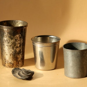Op de afbeelding: Drie metalen bekers van verschillende maten en texturen worden getoond. De hoogste beker heeft een gehamerde afwerking, de middelste beker is glad en glanzend en de kleinste heeft een ruwe, matte uitstraling. Een fossiel ligt ervoor.