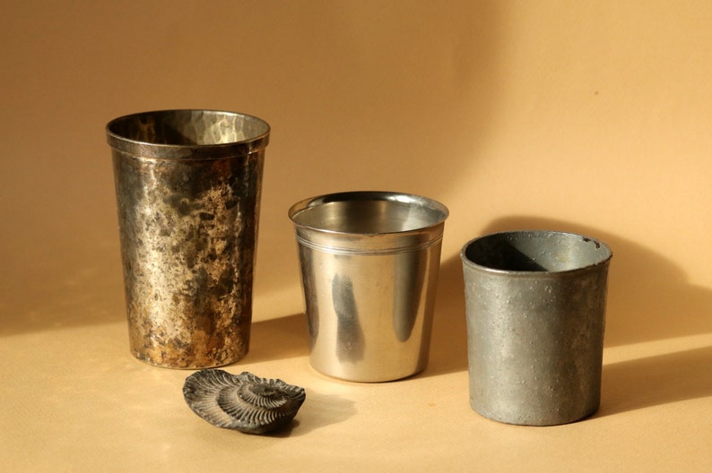 Op de afbeelding: Drie metalen bekers van verschillende maten en texturen worden getoond. De hoogste beker heeft een gehamerde afwerking, de middelste beker is glad en glanzend en de kleinste heeft een ruwe, matte uitstraling. Een fossiel ligt ervoor.
