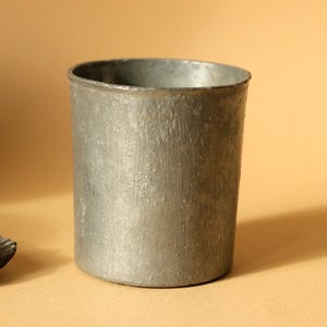 Op de afbeelding: Een cilindrische, grijze metalen beker met een getextureerd oppervlak. De beker heeft een licht uitlopende rand en een eenvoudig ontwerp. De beker is waarschijnlijk gemaakt van tin of een vergelijkbaar metaal.