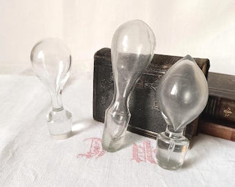 May include: Three clear glass bottle stoppers with rounded, bulbous tops and cylindrical bases. The stoppers vary in shape, with one featuring a pointed tip, while the others have smooth, rounded profiles. They are made of transparent glass.