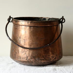 May include: A large, copper pot with a single iron handle. The pot has a rounded bottom and a slightly flared top. The copper has a patina and shows signs of age.