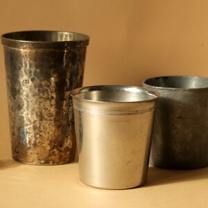 Op de afbeelding: Drie antieke metalen drinkbekers van verschillende formaten. De grootste beker heeft een gehamerde textuur, terwijl de kleinste een glad, gepolijst oppervlak heeft. De bekers zijn zilver en grijs, tegen een neutrale achtergrond.