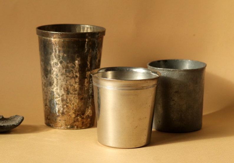 Op de afbeelding: Drie antieke metalen drinkbekers van verschillende formaten. De grootste beker heeft een gehamerde textuur, terwijl de kleinste een glad, gepolijst oppervlak heeft. De bekers zijn zilver en grijs, tegen een neutrale achtergrond.
