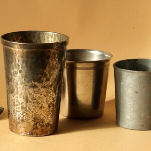 Op de afbeelding: Drie antieke metalen drinkbekers van verschillende maten en afwerkingen. De grootste beker heeft een getextureerde, gehamerde uitstraling met donkere patina. De andere twee bekers zijn kleiner, met een gladde, zilverkleurige afwerking. Een fossiel staat op de achtergrond.