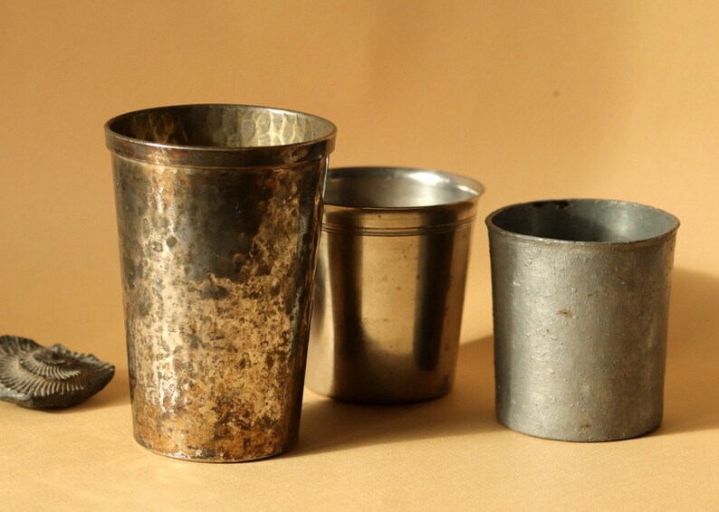Op de afbeelding: Drie antieke metalen drinkbekers van verschillende maten en afwerkingen. De grootste beker heeft een getextureerde, gehamerde uitstraling met donkere patina. De andere twee bekers zijn kleiner, met een gladde, zilverkleurige afwerking. Een fossiel staat op de achtergrond.