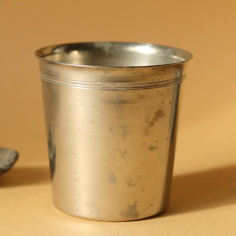 Op de afbeelding: Een vintage zilverkleurige metalen beker met een licht aangetaste afwerking. De beker heeft een eenvoudige, cilindrische vorm met een subtiele horizontale band aan de bovenkant. De achtergrond is een warme, neutrale tint.