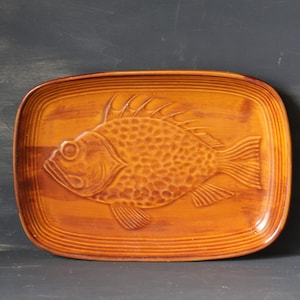 May include: A brown ceramic serving tray featuring a raised design of a fish. The tray is slightly rounded rectangular in shape and has a textured surface.