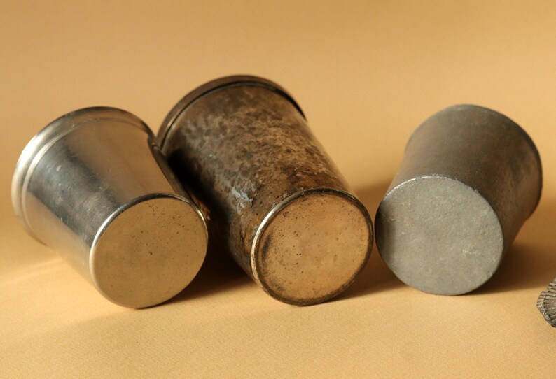 Op de afbeelding: Drie antieke metalen bekers in verschillende maten en afwerkingen. De bekers zijn gemaakt van zilver- en bronskleurige metalen. De beker links is zilverkleurig, de middelste beker heeft een gehamerde textuur en de rechter beker is donkergrijs.