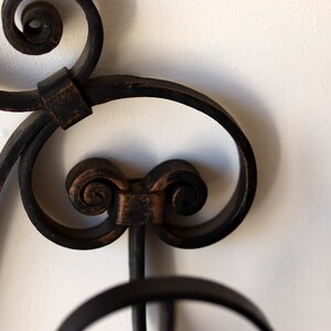 Op de afbeelding: Close-up van een zwarte metalen wanddecoratie met ingewikkelde krulwerk en een decoratief ontwerp. Het metaal is licht verweerd, waardoor het een vintage uitstraling heeft.