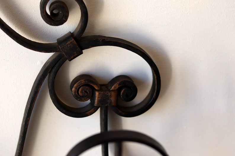 Op de afbeelding: Close-up van een zwarte metalen wanddecoratie met ingewikkelde krulwerk en een decoratief ontwerp. Het metaal is licht verweerd, waardoor het een vintage uitstraling heeft.