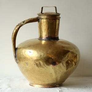 以下が含まれることがあります： 蓋と取っ手のある、大きなアンティークの真鍮製のピッチャー。ピッチャーは丸みを帯びたボディとハンマー仕上げになっています。