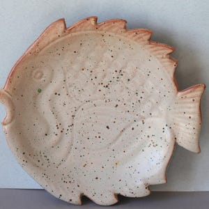 Peut inclure: Un plat en céramique blanche en forme de poisson avec des taches brunes et un motif en relief. Le plat est parfait pour servir des en-cas ou des amuse-gueules.