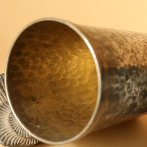 Op de afbeelding: Close-up van een gehamerde metalen beker met een getextureerd interieur. De beker heeft een zilveren rand en een donkere, verouderde buitenkant. Een fossiel is gedeeltelijk zichtbaar aan de linkerkant. De binnenkant van de beker heeft een gouden tint.