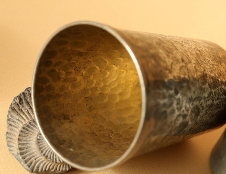 Op de afbeelding: Close-up van een gehamerde metalen beker met een getextureerd interieur. De beker heeft een zilveren rand en een donkere, verouderde buitenkant. Een fossiel is gedeeltelijk zichtbaar aan de linkerkant. De binnenkant van de beker heeft een gouden tint.