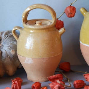 May include: Two ceramic crocks with lids, one yellow and one brown, with a white stripe. The crocks are sitting on a gray surface with dried orange flowers.