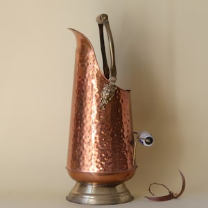 May include: A copper coal scuttle with a hammered texture, featuring a brass handle and decorative grape cluster. The scuttle has a white and blue ceramic handle and sits on a brass base.
