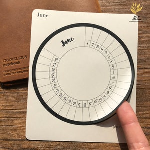 May include: A white circular sticker with a black border and a black circle in the center. The sticker is labeled "June" and has numbers 1 through 30 around the outer edge.