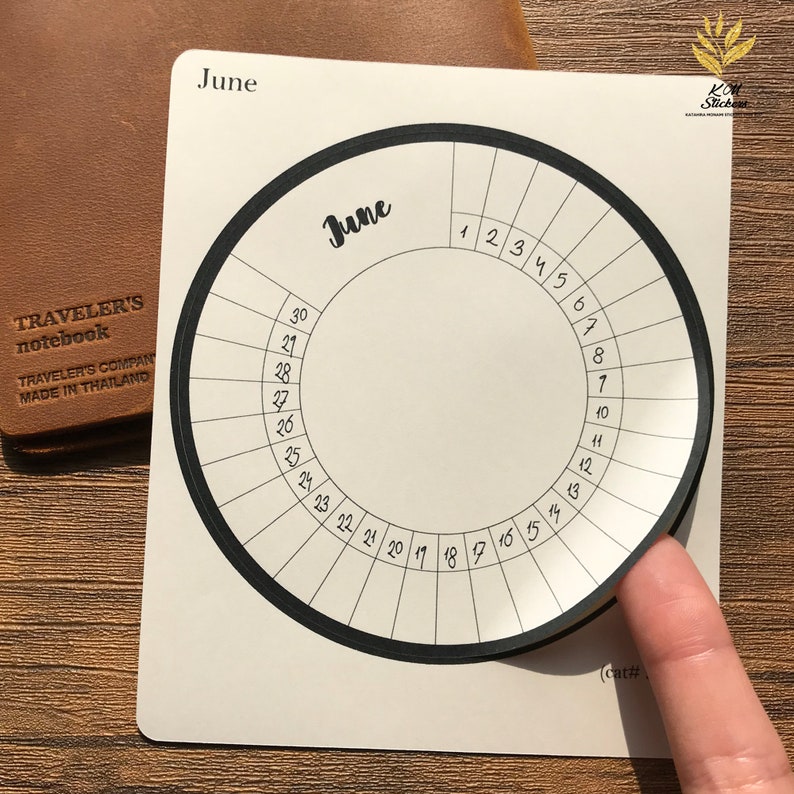 May include: A white circular sticker with a black border and a black circle in the center. The sticker is labeled "June" and has numbers 1 through 30 around the outer edge.