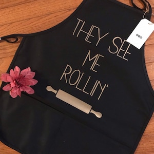 Peut inclure: Tablier noir avec une inscription dorée qui dit "They See Me Rollin'" avec un dessin de rouleau à pâtisserie. Le tablier a une grande poche sur le devant.
