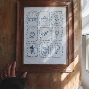 以下が含まれることがあります： 白い背景に青い刺繍が施された額装された刺繍作品。刺繍は、花、カップ、バスケット、ランプなど、さまざまなデザインの9つの正方形で構成されています。
