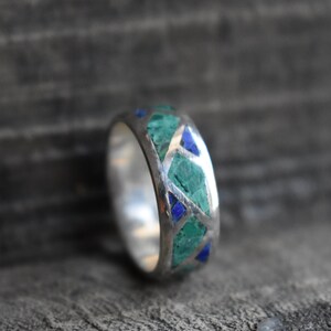 May include: A silver ring with a band of green and blue stones set in a geometric pattern.