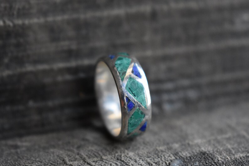 May include: A silver ring with a band of green and blue stones set in a geometric pattern.