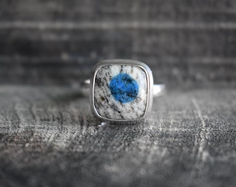 May include: A silver ring with a square blue and white stone set in the center.