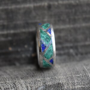 May include: A silver ring with a geometric pattern of green and blue stones.