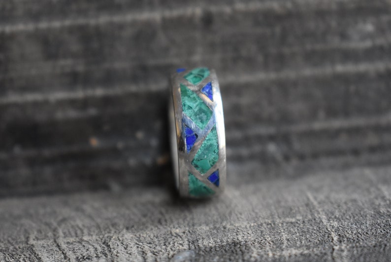 May include: A silver ring with a geometric pattern of green and blue stones.