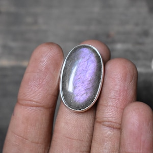 May include: Oval-shaped labradorite gemstone ring with a silver-colored setting. The gemstone displays a vibrant purple and blue iridescence. The ring is held in a hand, showcasing the polished stone's color and texture.