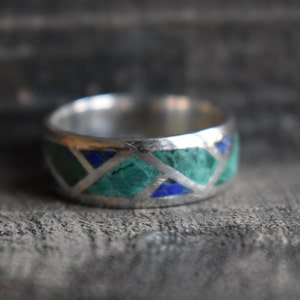 May include: A silver band ring with a geometric pattern of green and blue stones.