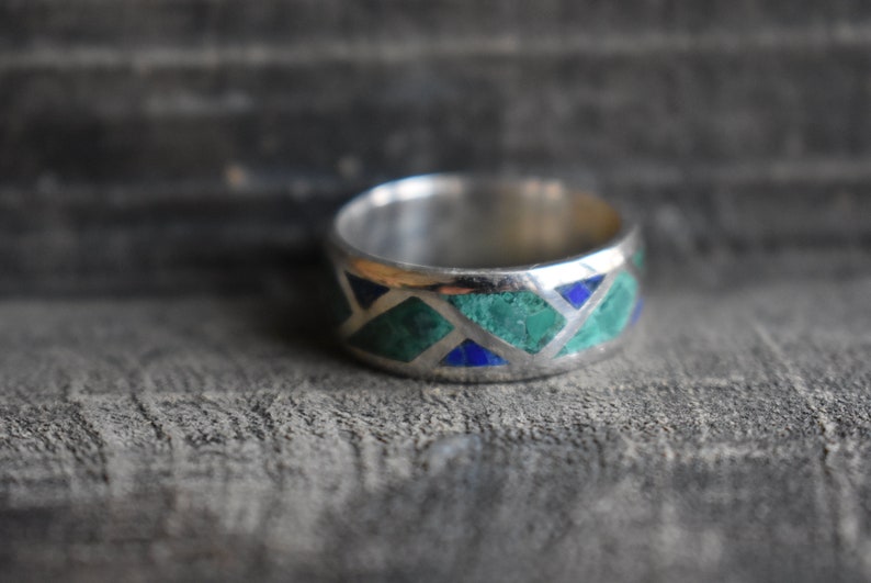 May include: A silver band ring with a geometric pattern of green and blue stones.