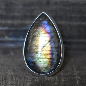 Puede incluir: Un anillo en forma de lágrima con una piedra preciosa de labradorita iridiscente engastada en una banda de plata.