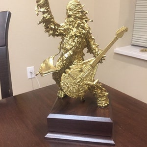 Puede incluir: Una figurita dorada de un Wookiee tocando una guitarra, de pie sobre una base de madera oscura.