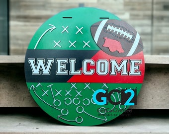 May include: A round, green wooden sign with a football theme. The word "WELCOME" is prominently displayed in white block letters. A football graphic and play diagrams are also featured. The sign also includes the text "GO 2".