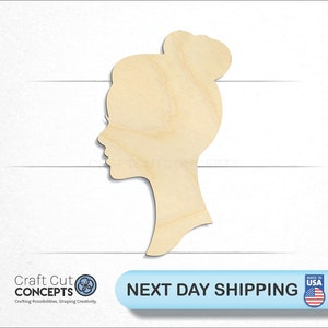 May include: A wooden cutout of a person's head in profile with a bun in their hair. The cutout is made of light brown wood and is approximately 5 inches tall.