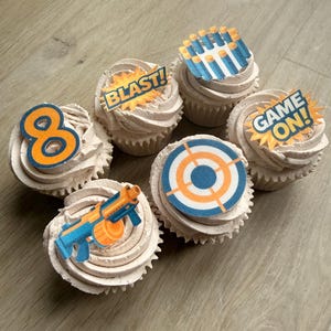 Könnte beinhalten: Sechs Cupcakes mit hellbraunem Frosting und thematischen Toppings. Die Toppings zeigen eine blaue und orangefarbene Zahl 8, eine Zielscheibe, eine Spielzeugpistole und die Sätze "BLAST!" und "GAME ON!".