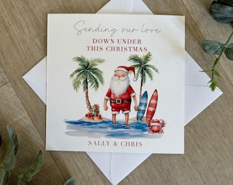 May include: A Christmas card featuring a cartoon Santa Claus wearing red shorts and a red and white Santa hat, standing on a beach with palm trees and a surfboard. The card says "Sending our love down under this Christmas" and "Sally & Chris".
