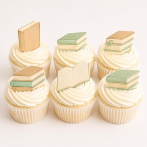 Puede incluir: Seis cupcakes con glaseado blanco y envoltorios de papel. Cada cupcake está decorado con un pequeño libro decorativo de papel marrón y verde. Un libro está abierto, los otros están cerrados.