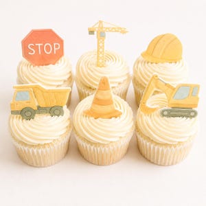 Puede incluir: Seis cupcakes con glaseado de vainilla y adornos con temática de construcción. Los adornos incluyen una señal de stop, una grúa, un casco, un camión volquete, un cono de tráfico y una excavadora.