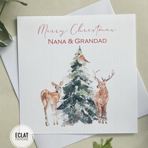 May include: A Christmas card with a watercolor illustration of a snowy forest scene. A Christmas tree with a robin perched on a branch is surrounded by two deer. The card reads "Merry Christmas Nana & Grandad".
