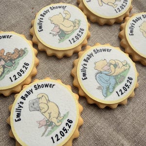 May include: A collection of round biscuits with a yellow, scalloped edge. Each biscuit features a white circular top with a Winnie the Pooh character illustration and the text "Emily's Baby Shower 12.05.26" in black. They are arranged on a textured, neutral-toned surface.