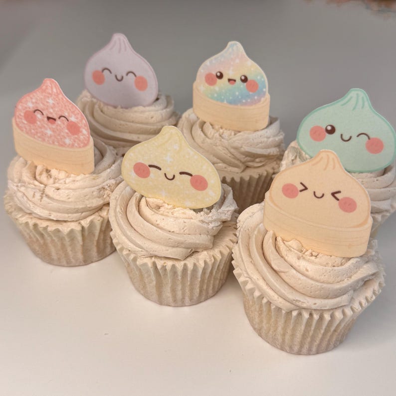 May include: Six cupcakes topped with beige frosting and decorative paper toppers. Each topper features a different pastel-colored, smiling dumpling character with rosy cheeks. The toppers include pink, lavender, rainbow, yellow, mint, and peach designs.