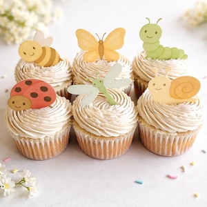 Puede incluir: Seis cupcakes con glaseado en espiral están cubiertos con decoraciones con temas de insectos. Se presentan una abeja, una mariposa, una mariquita, una libélula, una oruga y un caracol. Los cupcakes están dispuestos sobre una superficie blanca, con flores blancas en el fondo.