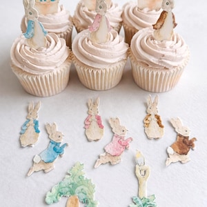 May include: Nine cupcakes with pale pink icing, each decorated with a watercolour bunny figure. Additional bunny cutouts, a carrot and a candle illustration are scattered around the cupcakes, creating an Easter theme.
