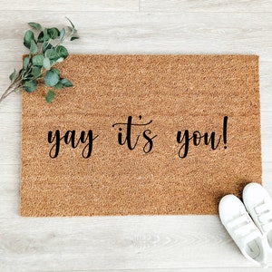 May include: A rectangular coir doormat with the phrase "yay it's you!" in black script. A sprig of greenery and a pair of white sneakers are placed near the mat. The mat is a natural brown color.